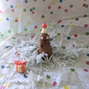 May include: A brown clay figurine of a person wearing a red and white party hat. The figurine is surrounded by white shredded paper and a small pink and white gift box.
