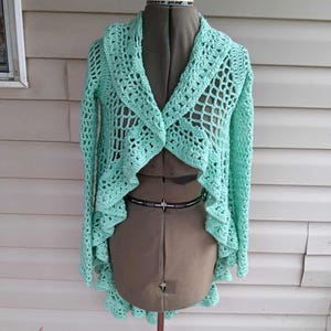 May include: A light turquoise crochet cardigan with long sleeves and a scalloped hem. The cardigan is displayed on a dress form. The crochet pattern features a mix of solid and openwork designs. The background is a neutral colour.