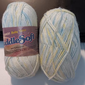 May include: Two skeins of Caron Baby Yarn in a pastel color palette of blue, yellow, and white. One skein has a label that reads "Caron Baby Yarn CuddleSoft". The yarn is soft and fluffy, ideal for baby projects.