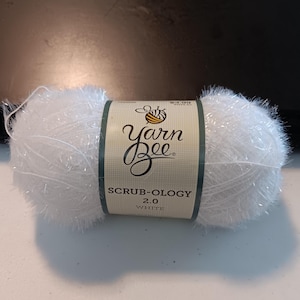Puede incluir: Ovillo de lana blanco Yarn Bee Scrub-ology 2.0. La etiqueta muestra el logotipo de Yarn Bee y los detalles del producto. La lana parece suave y esponjosa, ideal para proyectos de manualidades.