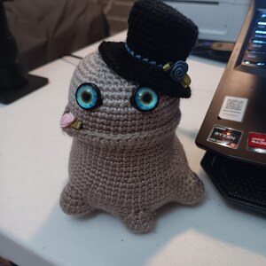 May include: A beige crocheted creature with large, bright blue eyes and a small pink flower in its mouth. It wears a black top hat adorned with a blue ribbon, a small blue rose, and a gold-colored accent piece on the side of the hat.