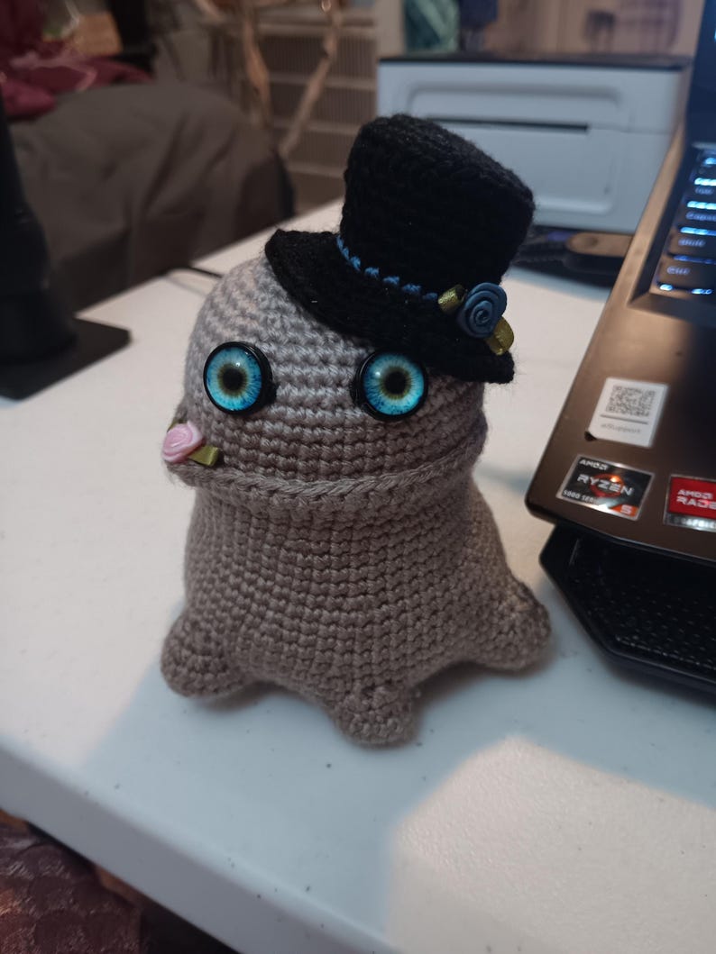 May include: A beige crocheted creature with large, bright blue eyes and a small pink flower in its mouth. It wears a black top hat adorned with a blue ribbon, a small blue rose, and a gold-colored accent piece on the side of the hat.