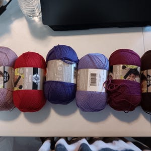 May include: Six skeins of yarn in various colors: mauve, red, deep blue, lavender, burgundy, and dark brown. Each skein has a label with a picture of a person wearing a hat. The yarn is arranged in a row on a white surface.