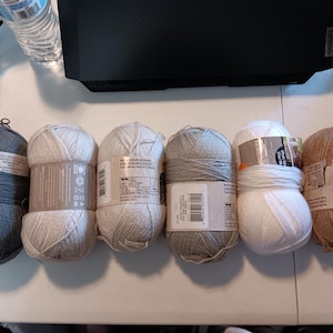 Puede incluir: Seis ovillos de lana en varios colores, incluyendo gris, blanco roto y marrón. Cada ovillo está envuelto con una etiqueta. La lana está dispuesta sobre una superficie blanca, adecuada para proyectos de tejido o ganchillo.