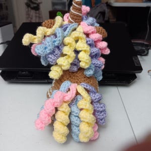May include: A handmade crochet unicorn with a brown body, a white and brown swirled horn, and colorful curly hair in shades of pink, yellow, blue, and purple. The unicorn is sitting on a black surface.