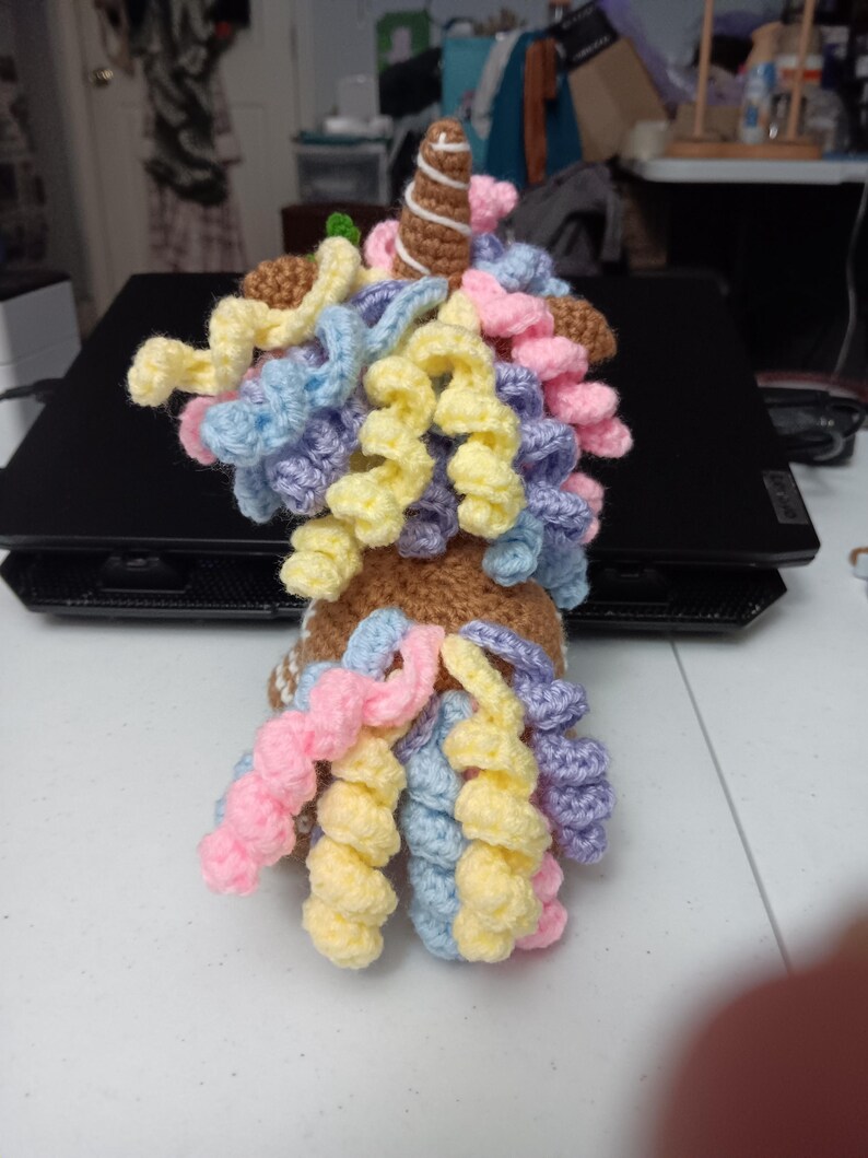 May include: A handmade crochet unicorn with a brown body, a white and brown swirled horn, and colorful curly hair in shades of pink, yellow, blue, and purple. The unicorn is sitting on a black surface.