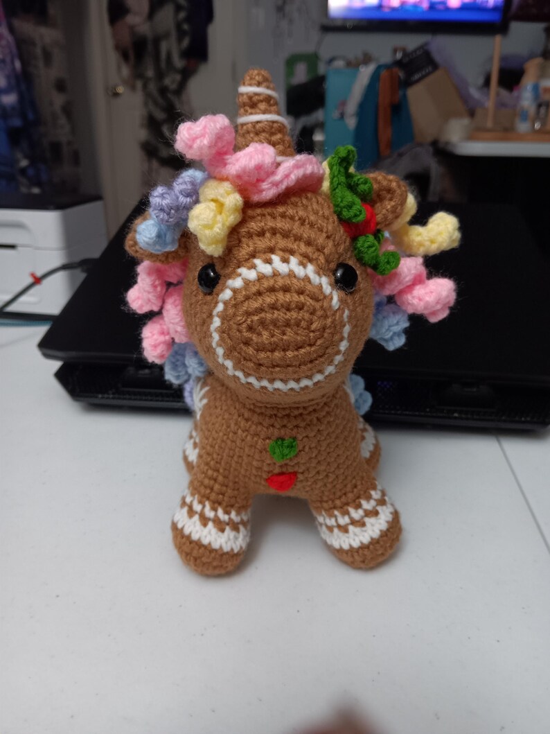 May include: A crocheted gingerbread unicorn with a brown body, white accents, and colorful yarn mane. The unicorn has a white horn, black eyes, and a small green and red heart-shaped design on its chest. The mane includes pink, yellow, and blue yarn.