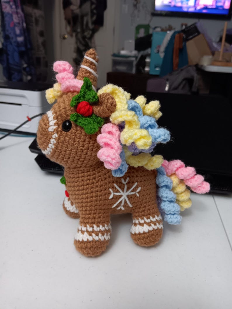 May include: A handmade brown crochet unicorn with a white snowflake design on its side. The unicorn has a white and brown striped horn, and a colorful mane and tail in shades of pink, yellow, and blue. It is decorated with a green holly and red berry.