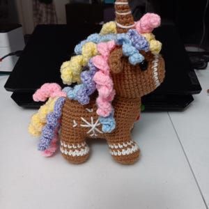 May include: A crocheted gingerbread unicorn with a brown body, white snowflake details, and a striped horn. Its mane is a mix of yellow, pink, and blue yarn. The unicorn has a whimsical, handmade appearance.
