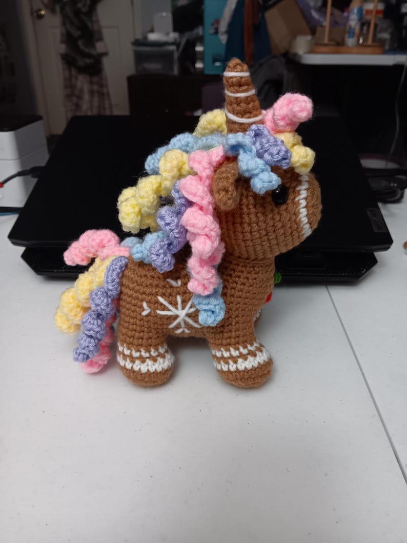 May include: A crocheted gingerbread unicorn with a brown body, white snowflake details, and a striped horn. Its mane is a mix of yellow, pink, and blue yarn. The unicorn has a whimsical, handmade appearance.