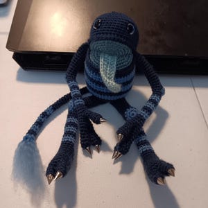 May include: A dark blue and light blue crocheted creature plush with a long tongue, striped limbs, a tail with a light blue tuft, and silver-colored pointed claws on its hands and feet. The plush has two dark eyes and a round, striped body.