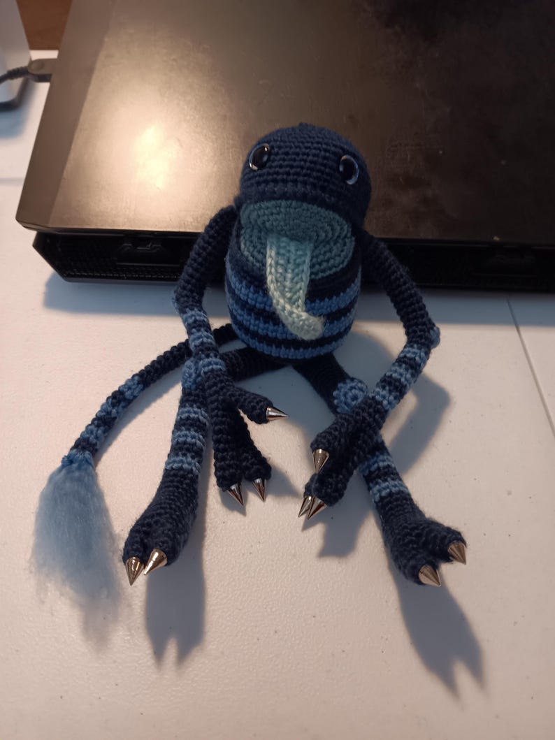 May include: A dark blue and light blue crocheted creature plush with a long tongue, striped limbs, a tail with a light blue tuft, and silver-colored pointed claws on its hands and feet. The plush has two dark eyes and a round, striped body.