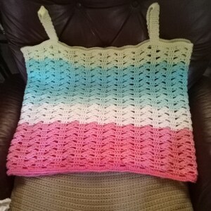 May include: A crocheted tank top with a multi-colored ombre design. The top is made with a light green, blue, white, and pink yarn. The top is laying on a brown crocheted surface.