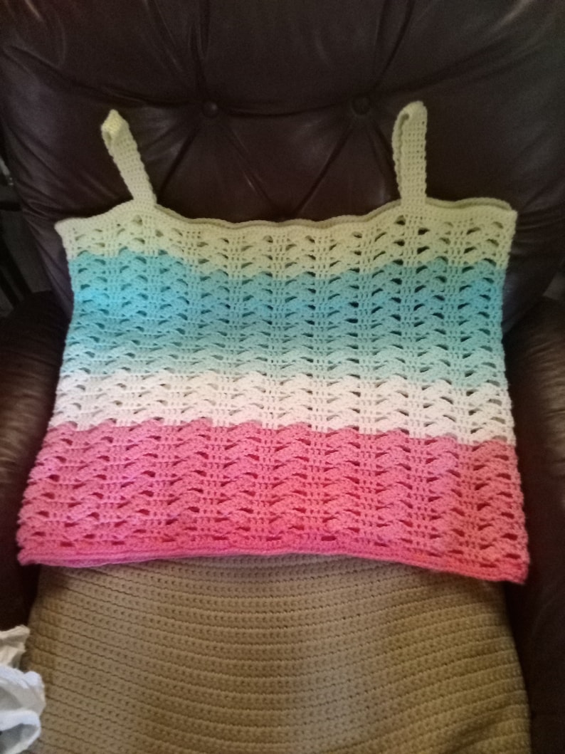 May include: A crocheted tank top with a multi-colored ombre design. The top is made with a light green, blue, white, and pink yarn. The top is laying on a brown crocheted surface.