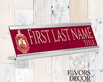 May include: A burgundy desk nameplate with the text "FIRST LAST NAME" and "TITLE" in a serif font. The nameplate has a silver frame and a Greek letter emblem on the left side. The background is a light wood pattern.