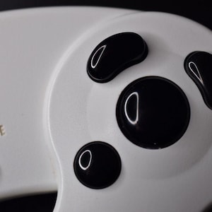 Puede incluir: Primer plano de un mando de Nintendo 64 blanco con botones negros. El mando tiene un logotipo de Nintendo Club rojo y blanco y el texto "Start/Pause".