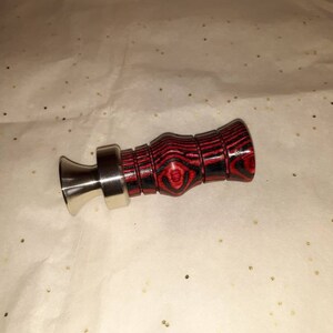 May include: A duck call with a stainless steel mouthpiece and a red and black wooden body. The wooden body has a textured, striped pattern. The duck call is designed for hunting and outdoor activities.