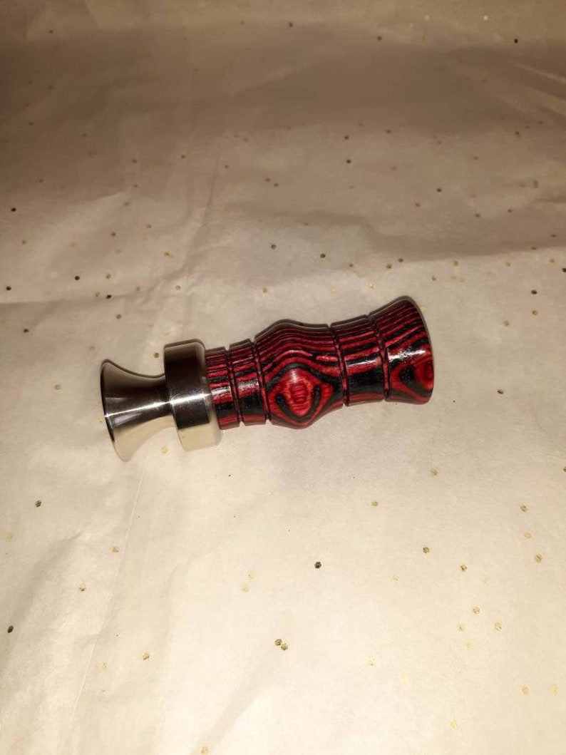 May include: A duck call with a stainless steel mouthpiece and a red and black wooden body. The wooden body has a textured, striped pattern. The duck call is designed for hunting and outdoor activities.