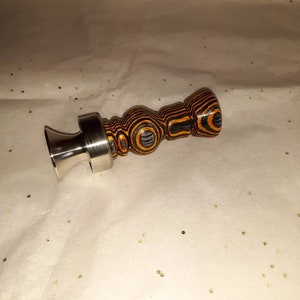 May include: A decorative bottle stopper with a stainless steel base and a wooden handle. The handle features a layered design with black and orange stripes. The stopper is designed to fit into a bottle opening.