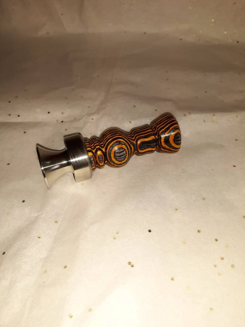 May include: A decorative bottle stopper with a stainless steel base and a wooden handle. The handle features a layered design with black and orange stripes. The stopper is designed to fit into a bottle opening.