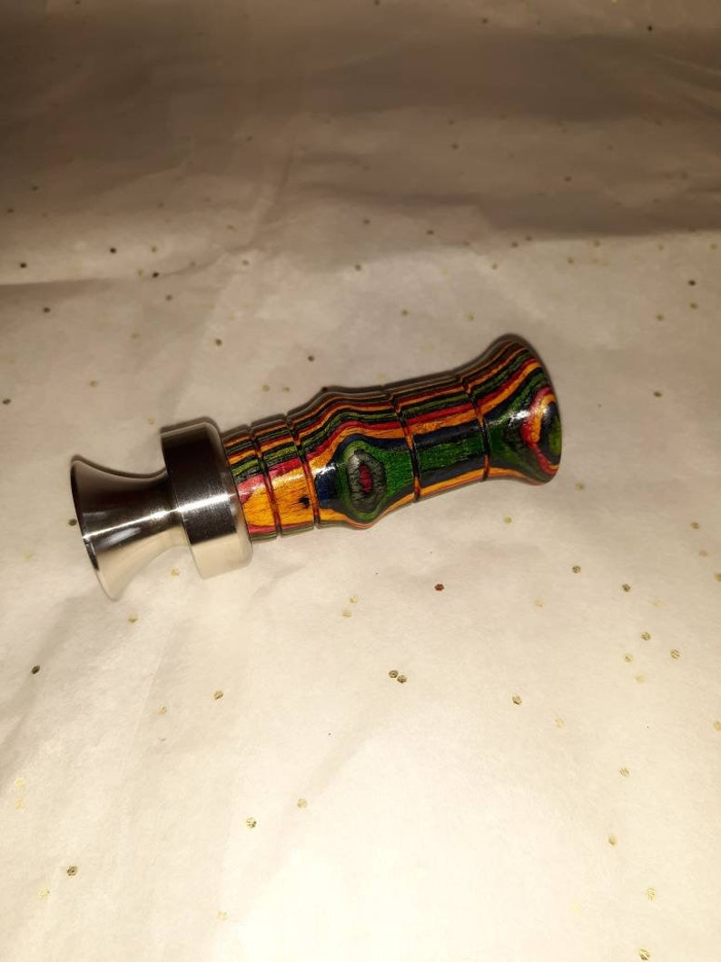 May include: A handcrafted bottle stopper with a stainless steel base and a colorful, layered wood handle. The handle features bands of red, green, yellow, and blue. The stopper is designed to fit into a bottle opening.