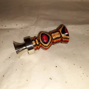 May include: A wooden duck call with a stainless steel mouthpiece. The duck call is made of layered wood in shades of brown, red, blue, and yellow. The call has a bulbous body and a flared mouthpiece.