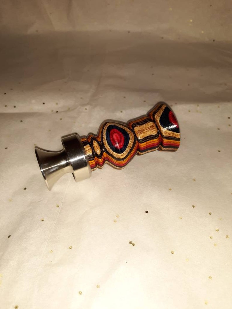 May include: A wooden duck call with a stainless steel mouthpiece. The duck call is made of layered wood in shades of brown, red, blue, and yellow. The call has a bulbous body and a flared mouthpiece.