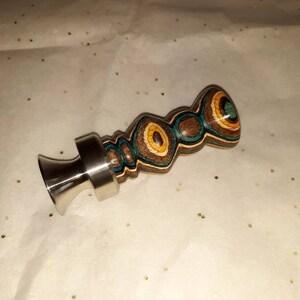 May include: A decorative bottle stopper with a stainless steel base and a wooden top. The wooden top features a layered design with brown, teal, yellow, and green circular patterns. The stopper is designed to fit into a bottle.