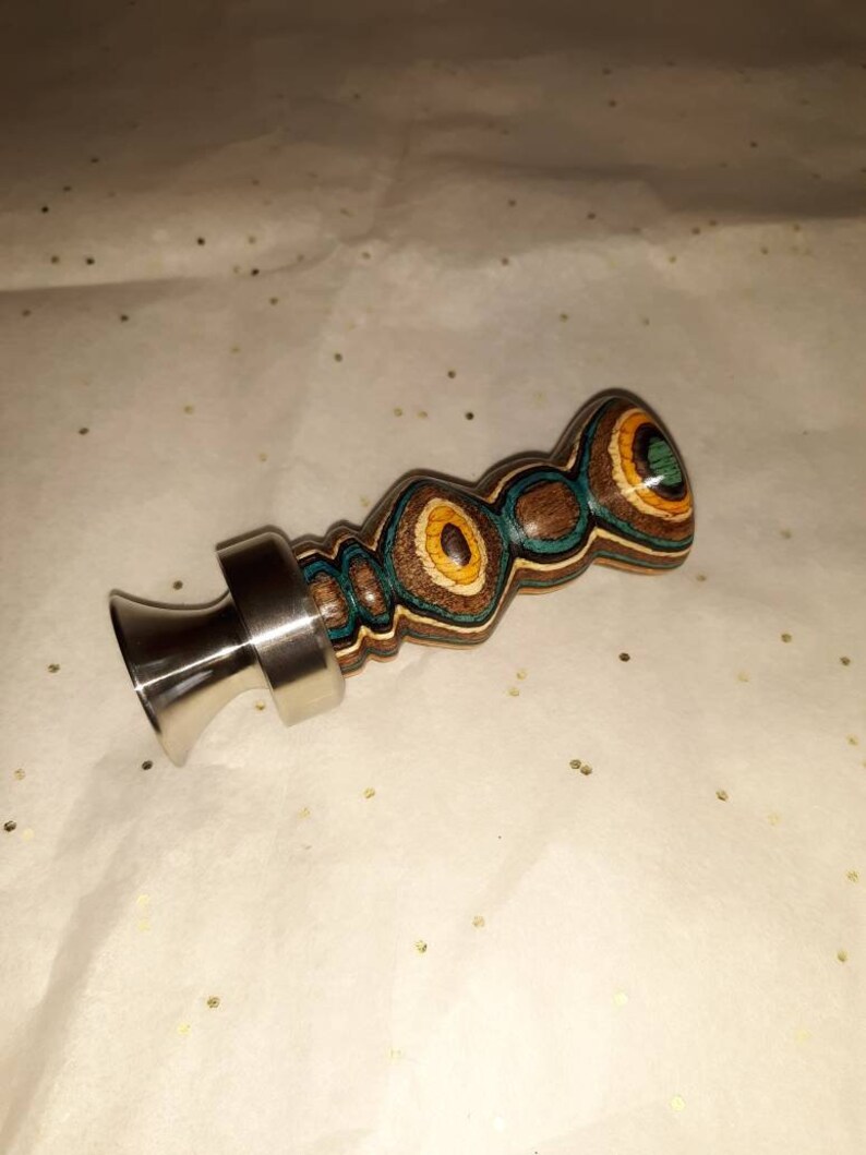 May include: A decorative bottle stopper with a stainless steel base and a wooden top. The wooden top features a layered design with brown, teal, yellow, and green circular patterns. The stopper is designed to fit into a bottle.