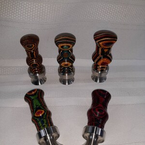 May include: Five decorative bottle stoppers with wooden tops in various colors and patterns, including brown, red, green, and blue. Each stopper has a silver-colored metal base. The stoppers are arranged on a white surface.