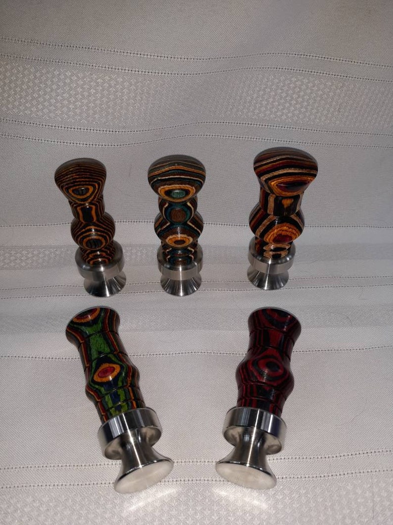 May include: Five decorative bottle stoppers with wooden tops in various colors and patterns, including brown, red, green, and blue. Each stopper has a silver-colored metal base. The stoppers are arranged on a white surface.