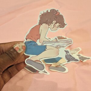 Op de afbeelding: Een sticker met een cartoonillustratie van een persoon die in een notitieboek schrijft, een rode top en een blauwe korte broek draagt. Er zijn ook twee kraaien afgebeeld. De sticker staat op een roze achtergrond.