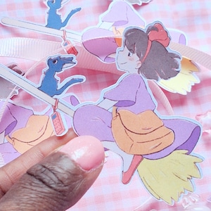 May include: Paper cutouts of a cartoon girl riding a broom, with a blue bird and a bag. The girl wears a purple dress and has a red bow in her hair. The background is a pink and white checkered pattern.