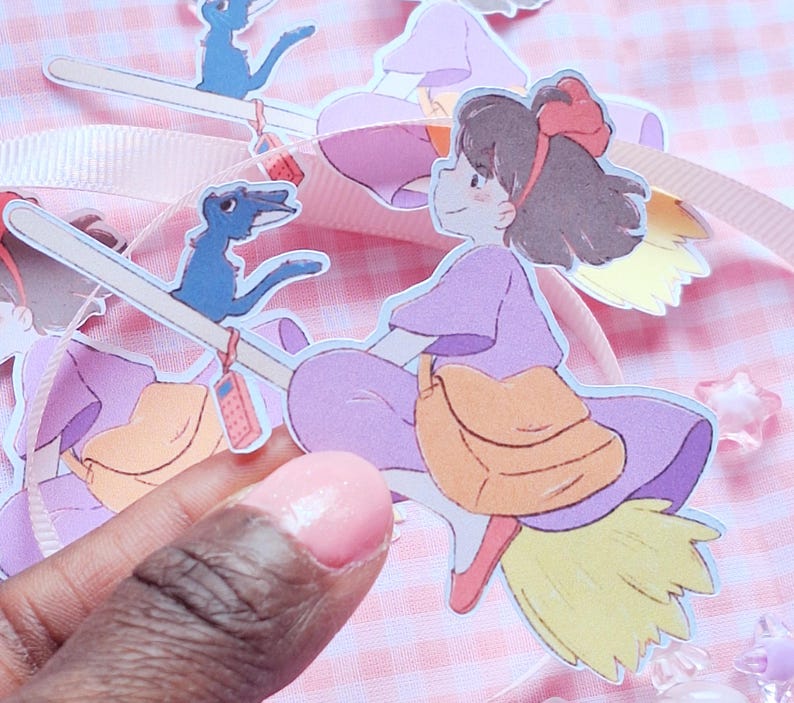 May include: Paper cutouts of a cartoon girl riding a broom, with a blue bird and a bag. The girl wears a purple dress and has a red bow in her hair. The background is a pink and white checkered pattern.
