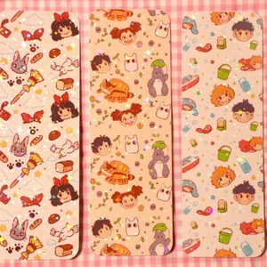 May include: Three rectangular bookmarks with colorful illustrations. Each bookmark features a different pattern of cartoon characters, food, and other objects. The background is a light peach color with glitter accents. The bookmarks are on a pink and white checkered surface.