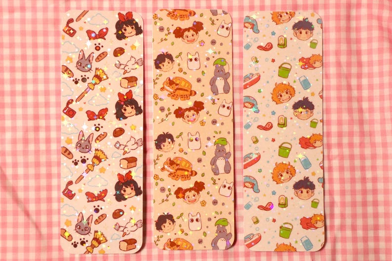 May include: Three rectangular bookmarks with colorful illustrations. Each bookmark features a different pattern of cartoon characters, food, and other objects. The background is a light peach color with glitter accents. The bookmarks are on a pink and white checkered surface.