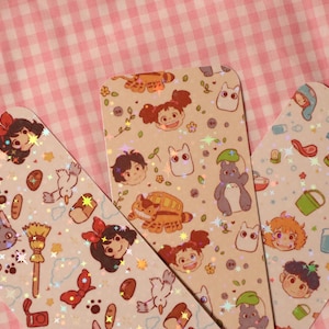 May include: Three decorative bookmarks with rounded corners, featuring colorful illustrations of anime characters and objects. The bookmarks have a light beige background with a holographic glitter effect. The background is a pink and white checkered pattern.
