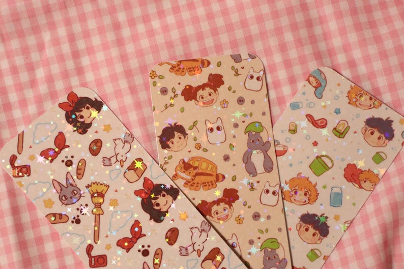 May include: Three decorative bookmarks with rounded corners, featuring colorful illustrations of anime characters and objects. The bookmarks have a light beige background with a holographic glitter effect. The background is a pink and white checkered pattern.