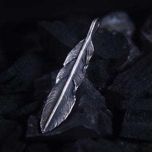 Peut inclure: Un pendentif en forme de plume argentée avec des détails complexes. Le pendentif est suspendu à une petite boucle et repose sur un lit de charbon de bois sombre et texturé. Le design de la plume est très détaillé, avec des barbes visibles et une tige centrale.