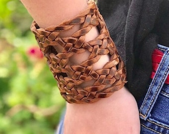 May include: A wide, brown leather cuff bracelet with a woven design. The bracelet is worn on a wrist, with the person wearing blue jeans and a black top. The background is blurred with green foliage.