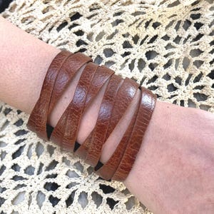 May include: A brown leather cuff bracelet with a woven design. The bracelet is made of multiple strips of leather that are woven together to create a unique and stylish look.
