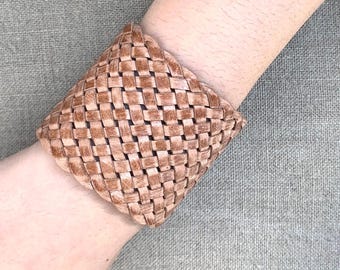May include: A woven leather cuff bracelet in a light brown color. The bracelet is made of braided leather and has a square shape.