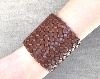 May include: A brown leather woven cuff bracelet. The bracelet is made of thick, braided leather and has a square shape.