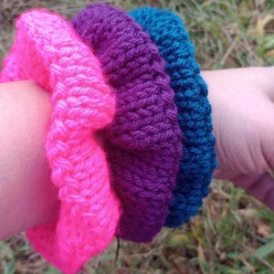 May include: Three knitted bracelets in pink, purple, and teal colors. The bracelets are stacked on top of each other on a person's wrist.