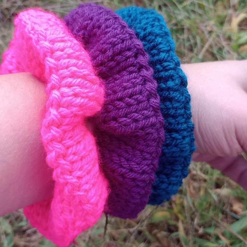May include: Three knitted bracelets in pink, purple, and teal colors. The bracelets are stacked on top of each other on a person's wrist.