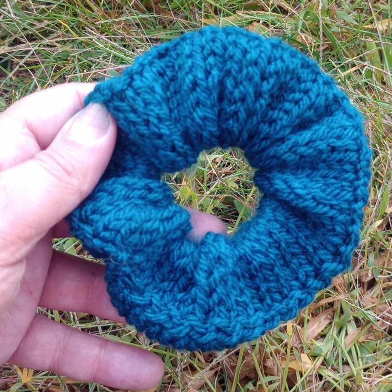 May include: A teal blue knitted hair scrunchie. The scrunchie is made of a soft, textured yarn and is designed to hold hair in place.