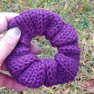 May include: A purple knitted hair scrunchie, made with a thick yarn, is held in a hand against a background of green grass.