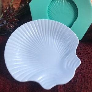 May include: A white ceramic seashell-shaped dish with a teal silicone mold of a seashell in the background.