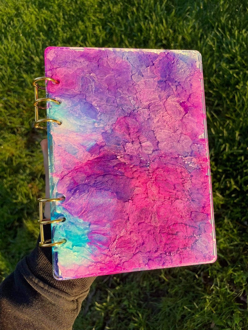 Może przedstawiać: Przezroczysty notes w oprawie żywicznej z kolorowym, abstrakcyjnym wzorem w odcieniach r&oacute;żowego, fioletowego i niebieskiego. Notes ma złote k&oacute;łka do wiązania.