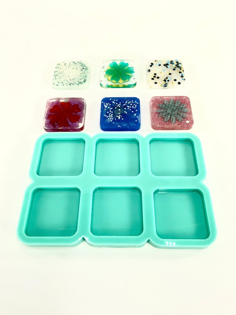 Op de afbeelding: Een turquoise siliconen mal met negen vierkante holtes voor het maken van harsambachten. Zes vierkante harsstukken met verschillende ontwerpen zijn te zien op een wit oppervlak.